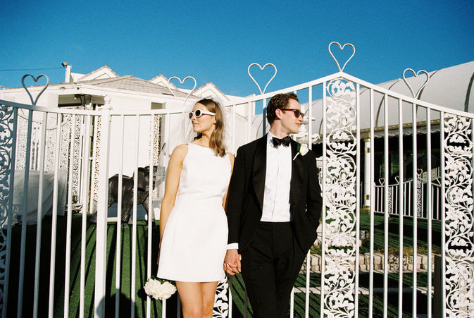 Bride and groom in sunglasses smile outside a white chapel under a clear blue sky. Veil and flowers; joyful wedding moment.