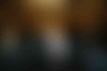 A couple in formal attire joyfully holds hands walking under glowing lights. Background: tiled wall, warm ambiance.