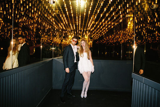 A couple in formal attire joyfully holds hands walking under glowing lights. Background: tiled wall, warm ambiance.