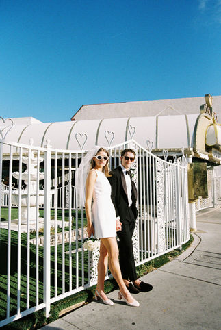 Bride and groom in sunglasses smile outside a white chapel under a clear blue sky. Veil and flowers; joyful wedding moment.