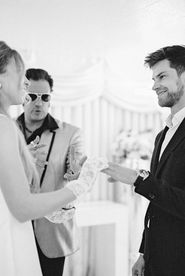 Bride in a white dress dances joyfully, groom claps, performer in gold jacket sings. Elegant floral backdrop with white drapery.