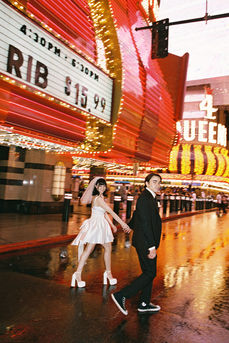 Couple under umbrella on wet street with neon hotel signs. Bride in white dress; groom in dark suit. Nighttime, city lights reflecting on pavement.
