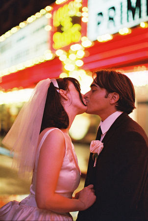 Couple under umbrella on wet street with neon hotel signs. Bride in white dress; groom in dark suit. Nighttime, city lights reflecting on pavement.
