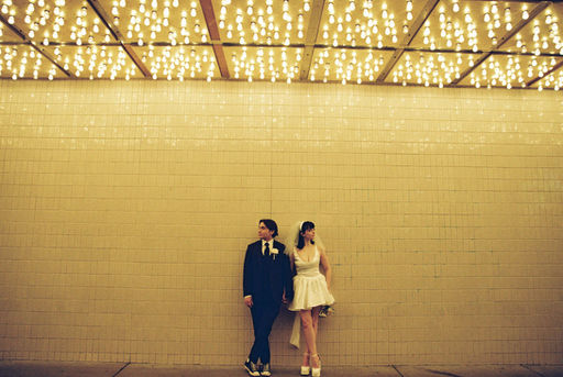 bride wearing white dress embraces groom while holding flowers and smiling underneath golden neon lights