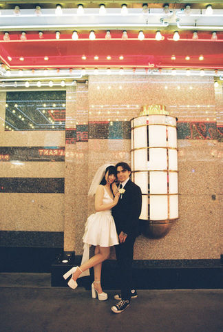 bride wearing white dress embraces groom while holding flowers and smiling underneath golden neon lights