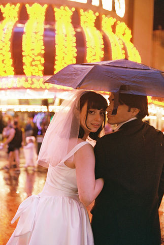 Couple under umbrella on wet street with neon hotel signs. Bride in white dress; groom in dark suit. Nighttime, city lights reflecting on pavement.