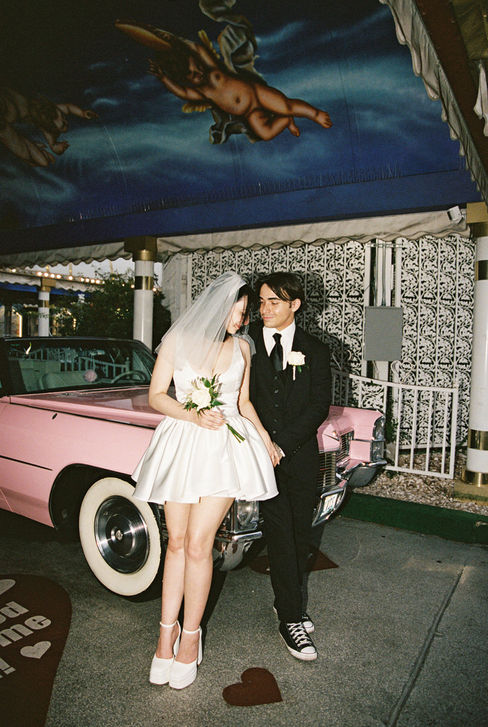 Bride and groom joyfully raise arms next to a pink convertible at a wedding chapel. Cupid mural above. Bride wears a white dress, groom in a suit.