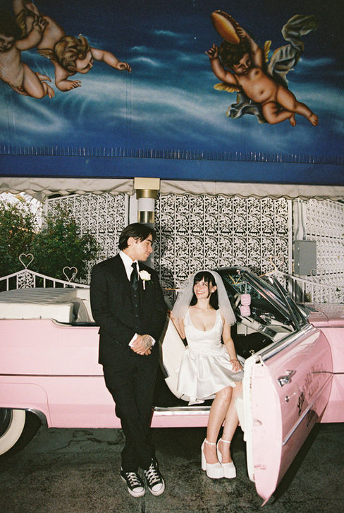 Bride and groom joyfully raise arms next to a pink convertible at a wedding chapel. Cupid mural above. Bride wears a white dress, groom in a suit.