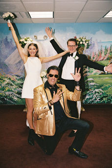 bride and groom stand at altar and smile with murals and chandeliers behind them at the little white chapel pose with elvis officiant on knee