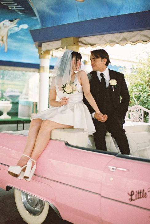Bride and groom joyfully raise arms next to a pink convertible at a wedding chapel. Cupid mural above. Bride wears a white dress, groom in a suit.