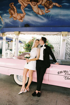 bride and groom wearing wedding attire kiss and hug while leaning against the side of a pink convertible with cupid mural overhead