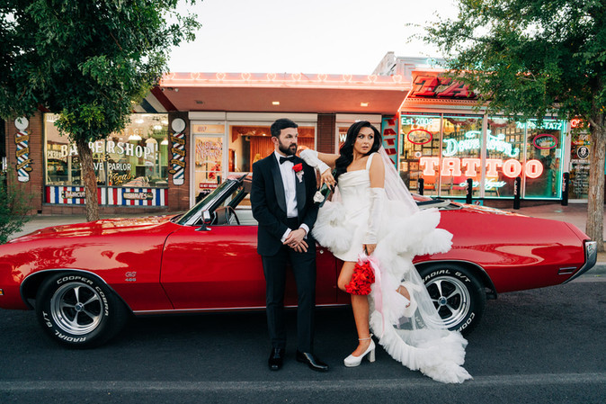 couple wearing wedding attire stand in front of a red convertible in downtown las vegas outside the sure thing chapel