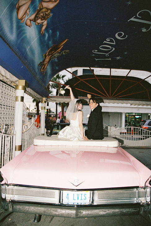 Bride and groom joyfully raise arms next to a pink convertible at a wedding chapel. Cupid mural above. Bride wears a white dress, groom in a suit.