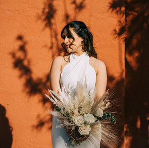 Bride poses in front of orange wall wearing a white halter top mini wedding dress, holding fall inspired boho bouquet with cream roses and pampas grass. Captured by Las Vegas elopement photographer brazen honey films.