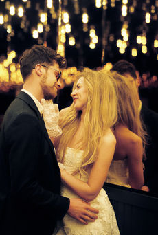 A couple in formal attire joyfully holds hands walking under glowing lights. Background: tiled wall, warm ambiance.