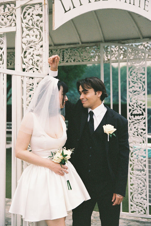 Bride and groom smile outside Little White Chapel, holding roses. Ornate white gate, green foliage in background. Joyous mood.