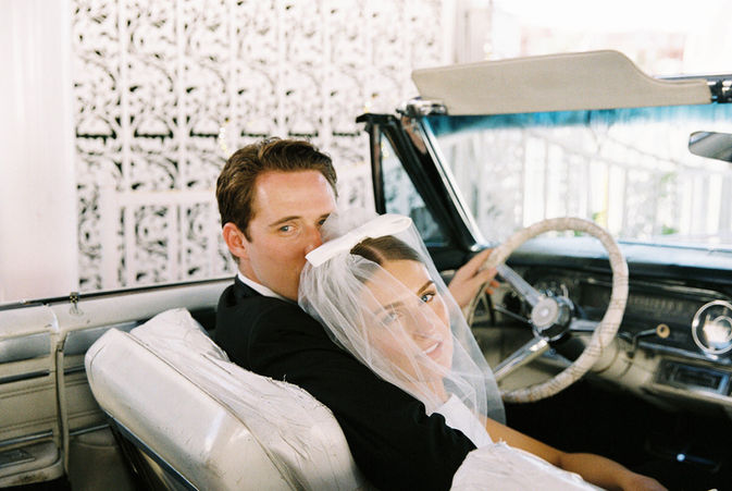 Bride kisses groom inside a pink convertible, holding rearview mirror. Both wear formal attire, creating a romantic and joyful mood.