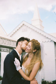 bride and groom embrace each other while he kisses her cheek, outside of a little white wedding chapel in las vegas; background: white chapel, blue skies
