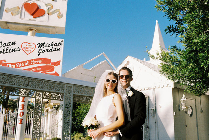 Bride and groom in sunglasses smile outside a white chapel under a clear blue sky. Veil and flowers; joyful wedding moment.