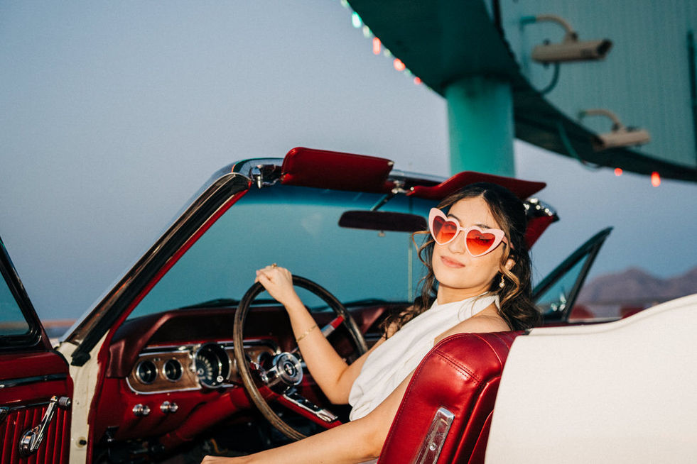 Bride sits in a convertible vintage mustang with red interior. Bride wears heart shaped sunglasses and wears halter top mini wedding dress. Best places to take wedding day pictures in Las Vegas. captured by las Vegas elopement photographer brazen honey films