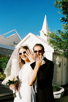 Bride and groom in sunglasses smile outside a white chapel under a clear blue sky. Veil and flowers; joyful wedding moment.