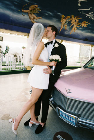 bride and groom wearing wedding attire kiss leaning against the hood of a pink cadillac