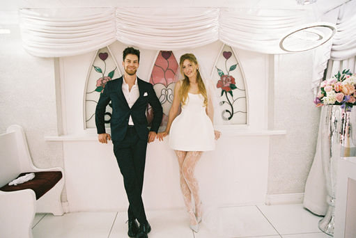 Bride in a white dress dances joyfully, groom claps, performer in gold jacket sings. Elegant floral backdrop with white drapery.