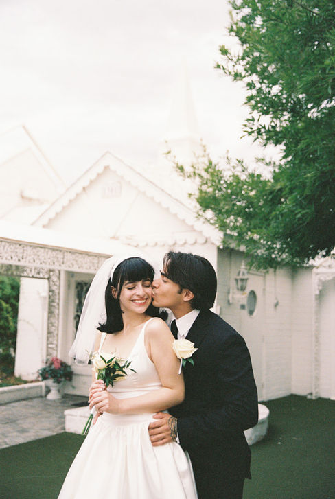 bride and groom wearing wedding attire take photos outside of the little white wedding chapel in las vegas groom kisses brides cheek