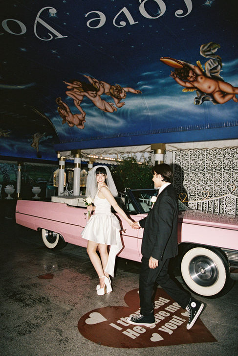 Bride and groom joyfully raise arms next to a pink convertible at a wedding chapel. Cupid mural above. Bride wears a white dress, groom in a suit.