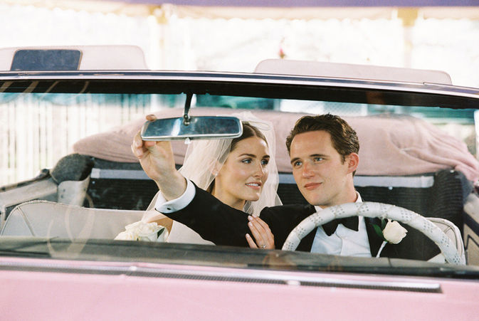 Bride kisses groom inside a pink convertible, holding rearview mirror. Both wear formal attire, creating a romantic and joyful mood.