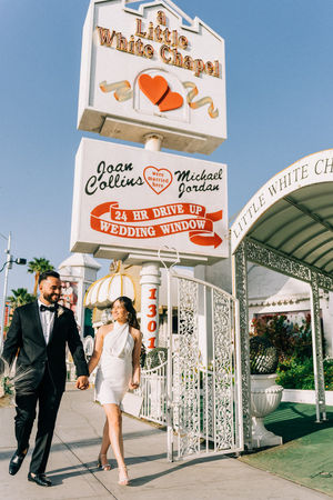 Bride and groom hold hands walking in front of the little white wedding chapel. Best places to take wedding day pictures in Las Vegas. captured by las Vegas elopement photographer brazen honey films