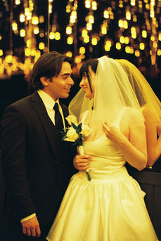 bride wearing white dress embraces groom while holding flowers and smiling underneath golden neon lights