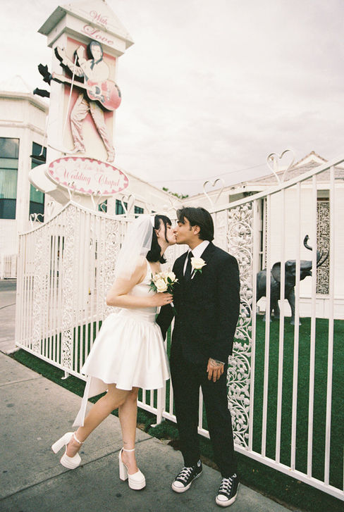 Bride and groom smile outside Little White Chapel, holding roses. Ornate white gate, green foliage in background. Joyous mood.