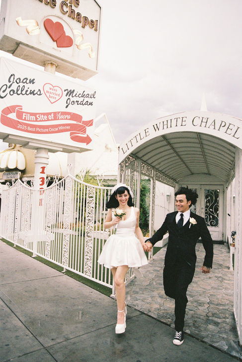 Bride and groom smile outside Little White Chapel, holding roses. Ornate white gate, green foliage in background. Joyous mood.
