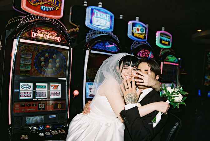 bride wearing white sits on groom lap in front of slot machines showing their ring hands and smiling
