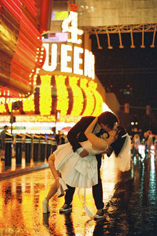 Couple under umbrella on wet street with neon hotel signs. Bride in white dress; groom in dark suit. Nighttime, city lights reflecting on pavement.