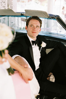 groom wearing black suit sits in a convertible and smiles