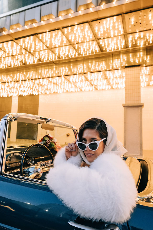 bride wearing white fur coat, vintage sunglasses and head scarf poses for photos in a blue convertible thunderbird. captured by las vegas wedding photographer brazen honey films