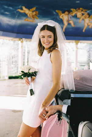 bride wearing short wedding dress and veil with vow leans against a pink convertible and smiles