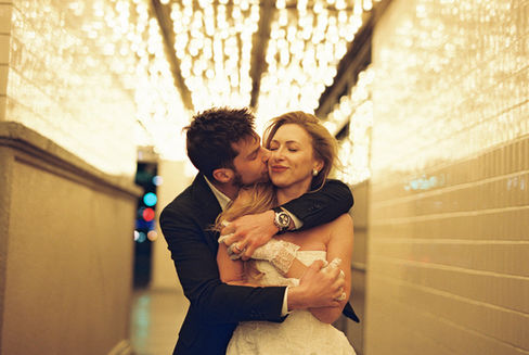 A couple in formal attire joyfully holds hands walking under glowing lights. Background: tiled wall, warm ambiance.