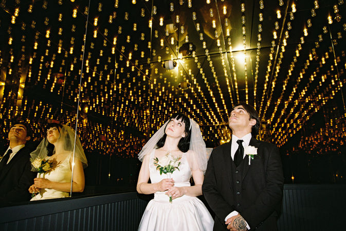 bride wearing white dress embraces groom while holding flowers and smiling underneath golden neon lights