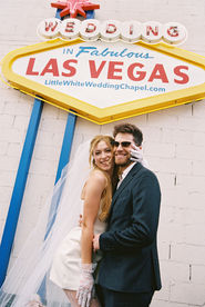 Bride in a white dress dances joyfully, groom claps, performer in gold jacket sings. Elegant floral backdrop with white drapery.