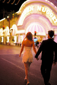 A couple in formal attire joyfully holds hands walking under glowing lights.