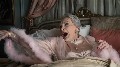 Poster showing an elderly woman in a pale pink robe and fur-trimmed sleeves sitting up in bed mid-shout, arm outstretched, in an ornate bedroom setting.