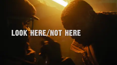 Warm, amber-lit image of two young men facing each other at close range in an intimate, low-lit setting. Their faces are partially in shadow, and the centred white title reads “Look Here / Not Here.”