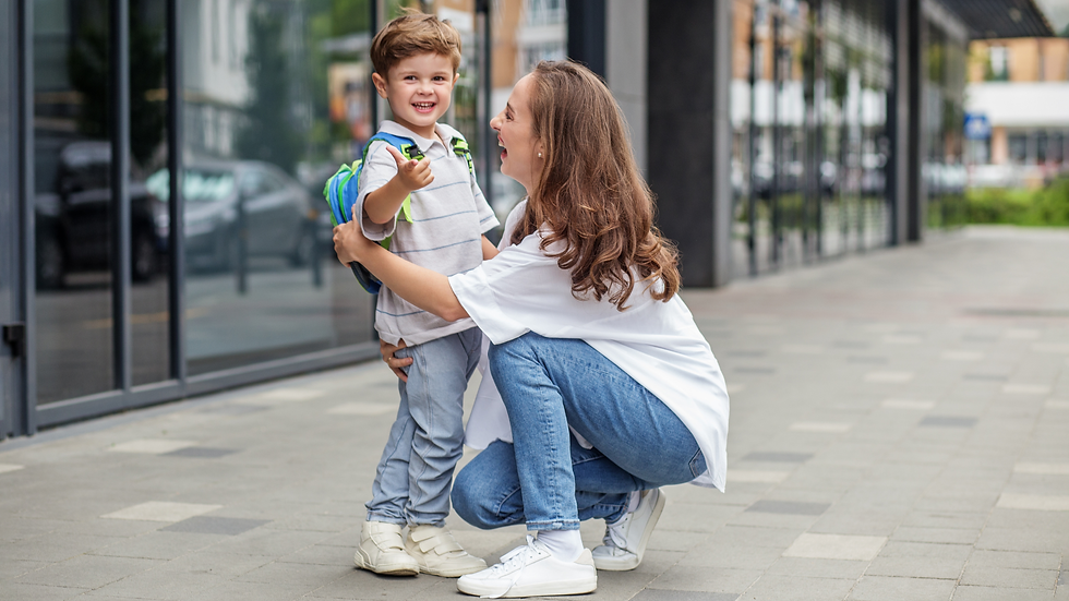 Mãe e filho sorrindo na calçada em frente a um prédio com portas envidraçadas, ambos vestem calça jeans, camiseta e tênis branco.