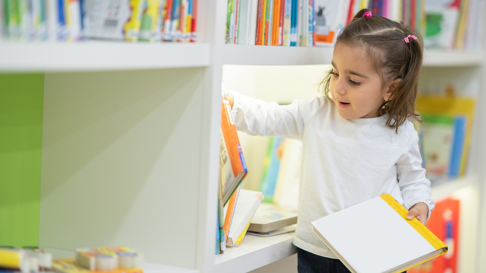 Criança com caderno na mão pegando um livro de uma prateleira branca, nichos com mais livros expostos.