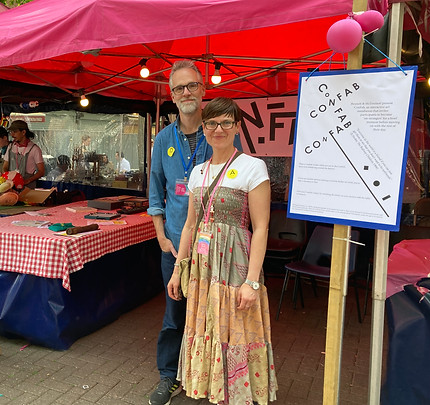 A white man dressed in a denim shirt standing behind a white woman in a white t-shirt and colourful dress. They are smiling.