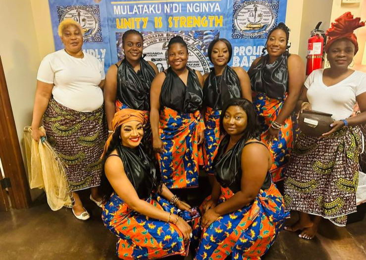 Group of women in colorful skirts and black tops, smiling, with the text 'Mulataku N'di Nginya Unity is Strength' behind them