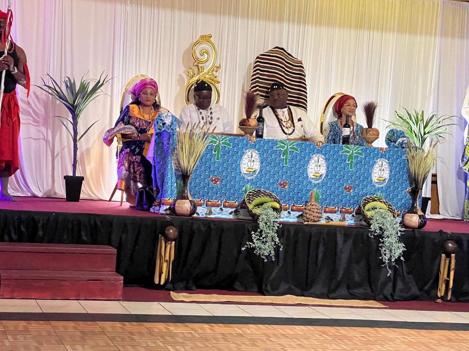 Four people seated at a table, with a man in traditional attire and a woman in a pink headwrap, on a stage with a decorated tablecloth and a palm tree
